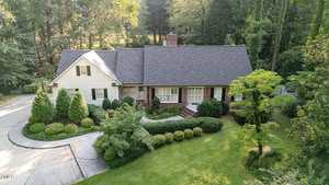 The Henderson home at 515 S Woodland Road is a two-story suburban house with a gabled roof and brick siding, surrounded by manicured bushes, trees, and a curved driveway leading to the entrance in a wooded area.