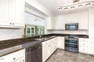 Modern kitchen with white cabinets, dark granite countertops, stainless steel appliances, a double sink, tile backsplash, and gray tile flooring. Large pass-through window opens to a bright living area with large windows.