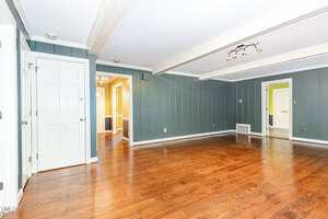 A spacious empty room with green paneled walls, hardwood floors, white trim, ceiling beams, and multiple doorways leading to adjoining rooms. The ceiling has a modern light fixture.