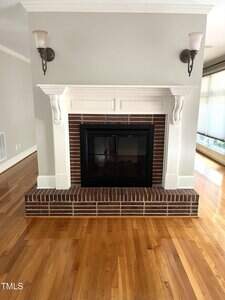 A brick fireplace with a white wood mantel is centered in the room. The fireplace has a black framed glass door. There are two wall-mounted light fixtures above the mantel, and the floor is hardwood.