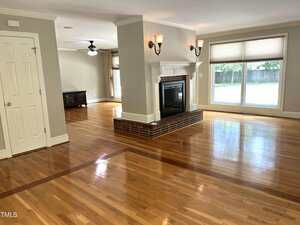 A spacious living area featuring polished hardwood floors, a central double-sided brick fireplace, large windows with blinds, and neutral-colored walls. Adjacent room visible with ceiling fan and TV stand.
