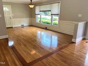 An empty room with light hardwood floors, a darker wood border, large front windows, white trim, and a hanging light fixture. The front door is visible on the left, and sunlight brightens the space.
