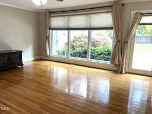 Empty room with light wood flooring, a large window with beige curtains, and a door to the right. Sunlight illuminates the room, and greenery is visible outside. A dark cabinet sits in the left corner.