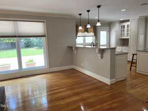 A bright, empty room with hardwood floors, a large window overlooking a backyard, and an open kitchen with white cabinets, granite countertops, pendant lights, and a breakfast bar with decorative supports.
