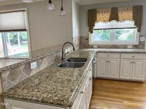 A kitchen with granite countertops, white cabinets, a double sink with a gooseneck faucet, tile backsplash, light wood flooring, and a window with a decorative valance overlooking a yard.