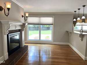 A living room with polished hardwood floors, a brick fireplace with white trim, wall-mounted light fixtures, a large window with blinds, and a kitchen counter with hanging pendant lights.