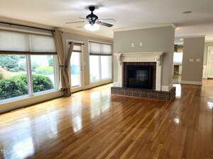 A spacious, empty living room with hardwood floors, large windows with beige curtains, a ceiling fan with lights, and a fireplace with a white mantel. Natural light fills the room, and there's an opening to a kitchen area.