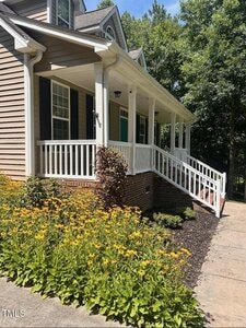 A one-story house with beige siding, white trim, and a front porch with railings. Yellow flowers and green shrubs line the walkway leading to the steps and front door. Trees are visible in the background.