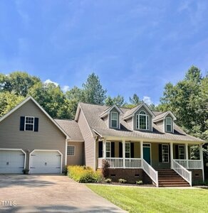 A tan, two-story house with a front porch, three dormer windows, white trim, and a two-car garage sits on a sunny day, surrounded by green trees and a well-kept lawn with yellow flowers by the driveway.