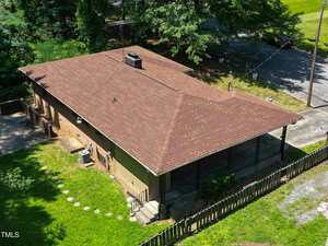 Aerial view of a single-story brick house with a brown hip roof, a covered front porch, fenced yard, and concrete driveway, surrounded by trees and grassy areas near a street.