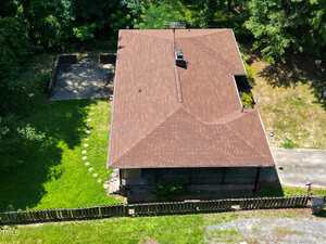 Aerial view of a single-story house with a red-brown roof, surrounded by grass, trees, and a wooden fence. A paved driveway and a patio area are visible beside the house.