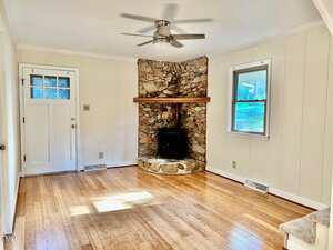 A bright living room with light wood floors, white paneled walls, a white door with a window, a single window, and a stone corner fireplace with a wood stove. A ceiling fan is mounted in the center.