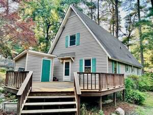 A light gray A-frame house with green-trimmed windows and a wooden deck is surrounded by trees and greenery. The house has a steep roof, an attached shed, and steps leading up to the front entrance.