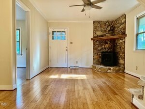 A bright living room with light-colored walls, wood flooring, a ceiling fan, a white front door with glass panes, and a corner stone fireplace with a wood mantle. Sunlight enters through two windows.