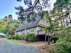 A two-story house with a steep roof, light-colored siding, and a wooden porch is surrounded by trees and shrubs. A gravel driveway leads to the entrance, and the yard is partially shaded.