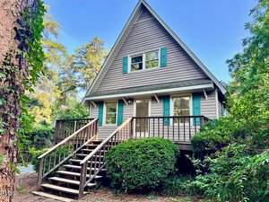 A gray A-frame house with teal shutters at 612 Williamsboro Street, Oxford, features a wooden porch and stairs leading to the entrance. Green shrubs and trees surround the home, all shaded by tall pines.