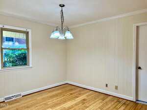 An empty room with light-colored wood flooring, cream-colored paneled walls, a window showing greenery outside, a white door, and a ceiling light fixture with five bulbs.