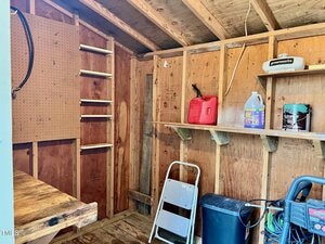 Interior of a wooden shed with unfinished walls, a small workbench, pegboard, shelves holding a gas can, liquid container, and cleaner, a white step ladder, and a black storage bin on the floor.