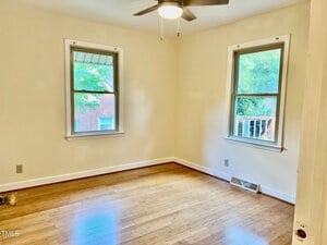 A small, empty room with light-colored walls, two windows, a ceiling fan with light, hardwood floors, and visible electrical outlets. Sunlight enters through the windows, showing trees and a brick building outside.