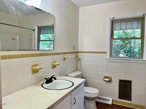 A bathroom with a white sink, black faucet, countertop, toilet, and a wall-mounted mirror. The walls are partially tiled with beige accents. Two windows with outside views are visible, along with a wall vent and a towel holder.