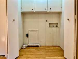 An empty laundry closet with white cabinets above, hook-ups for a washer and dryer, a flexible vent hose on the floor, wood flooring, and white doors open to reveal the space.