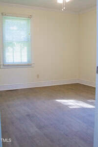 An empty room with light-colored walls, a window with blinds on the left wall, wood-style flooring, and a white ceiling with a ceiling fan partially visible at the top.