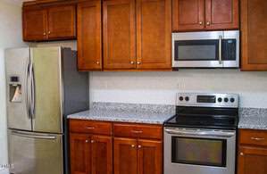 A kitchen with wooden cabinets, stainless steel refrigerator, built-in microwave, electric stove, and granite countertops. The backsplash is white with a subtle pattern. The kitchen appears clean and organized.