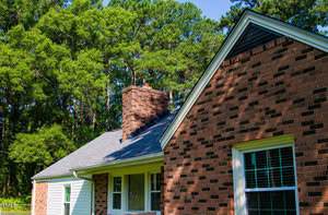 A close-up view of the exterior of a brick house with white trim, a brick chimney, several windows, and a background of tall green trees under a blue sky.