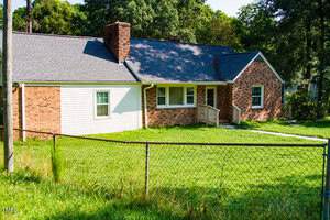 Single-story brick house with white siding on one side, a brick chimney, and a small porch with a railing. The yard is grassy and enclosed by a chain-link fence; trees are in the background.