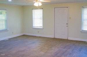 An empty room with light-colored walls, a wooden floor, three windows with blinds, and a white door. A ceiling fan with lights is mounted above. The room is well-lit with natural light.
