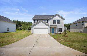 A two-story suburban house with gray siding, a blue front door, attached two-car garage, and concrete driveway. The front yard has grass, a small tree, and some potted plants by the entrance.