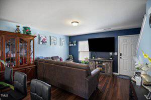 A living room with a brown sectional sofa, dark wood floors, a large wall-mounted TV, and a wooden display cabinet. The walls are blue and white, decorated with floral artwork and small shelves with decor.