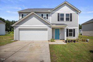 Two-story suburban house with gray siding and white trim, a double garage, a light blue front door, and a few small potted plants by the entrance. The driveway and lawn are visible in front.