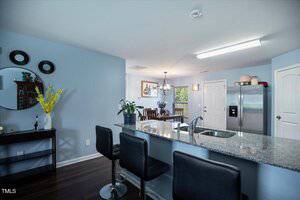 A modern kitchen with blue walls, dark wood floors, a granite island with three barstools, stainless steel appliances, and a dining area visible in the background near a window.