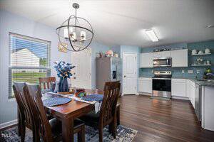 A modern kitchen and dining area with white cabinets, stainless steel appliances, blue backsplash, a wooden dining table set for four, a chandelier above, and a window showing a green lawn and a white house outside.