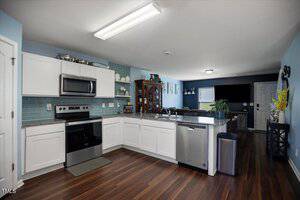 Modern kitchen with white cabinets, stainless steel appliances, blue tile backsplash, granite countertops, and dark wood flooring. The open layout leads to a living area with a TV, dining set, and large window.
