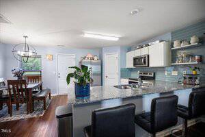 A modern kitchen with stainless steel appliances, blue backsplash, white cabinets, and a granite counter with three black barstools. A dining area with a wooden table and chairs is visible in the background.
