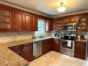 Kitchen with wooden cabinets, granite countertops, stainless steel appliances, a double sink under a window, beige tile backsplash, under-cabinet lighting, and a ceiling light fixture.