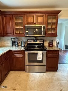 A kitchen features brown wooden cabinets, a granite countertop, stainless steel electric stove and oven, a mounted microwave, coffee maker, toaster, knife block, and beige tile floor and backsplash.