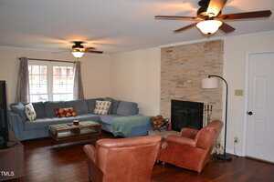 A living room with wood floors, a gray sectional sofa with cushions, two brown leather chairs, a coffee table, a stone fireplace with a log holder, two ceiling fans, and a floor lamp near the door.