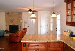 A view from a kitchen with granite countertops and wooden cabinets, looking into a living room with a stone fireplace, brown leather recliner, ceiling fan, and two white doors on the back wall.