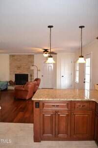 Interior view of a living room and kitchen area. The kitchen has brown cabinets and granite countertops, while the living room includes a stone fireplace, brown leather chair, hardwood floors, and hanging lights.
