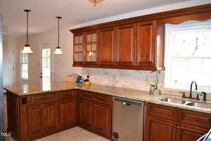 A kitchen with wooden cabinets, granite countertops, a tile backsplash, stainless steel dishwasher, double sink under a window, pendant lights, and a few small items on the counter.