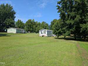 A single-story manufactured home sits on a large grassy lawn with trees in the background and another similar home to the left; the sky is clear and blue.