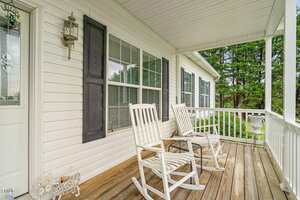 A white wooden front porch with two rocking chairs, a metal decorative pig, and a potted plant. The house has white siding, black shutters, and large windows. Trees and greenery are visible in the background.