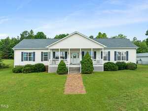 This charming single-story white house at 9633 Grassy Creek Road features a gray roof, front porch, and steps leading to a brick pathway. Green shrubs line the front lawn, with trees in the background under a clear Bullock blue sky.