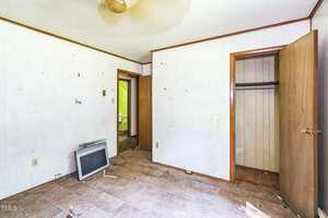 A mostly empty room with white paneled walls, tan ceiling, and brown trim. The floor is bare with some debris. An old TV lies on the floor, the closet door is open, and a second door leads to another room.