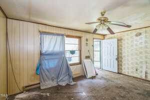 A small, cluttered room with wood panel walls, a ceiling fan, a window partially covered by a curtain and debris, a damaged screen, an open door, and stained carpeting. Walls show peeling wallpaper and dirt.