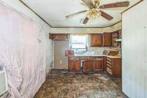 A worn kitchen with broken cabinets, dirty floors, a ceiling fan with missing blades, and stained walls. A pink sheet hangs on the left side. Natural light enters through a window above the sink.