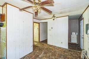 A room with white wood-paneled walls, a ceiling fan with four blades, brown tiled floor, a hanging light fixture, visible dirt and wear on surfaces, and doorways leading to other areas.